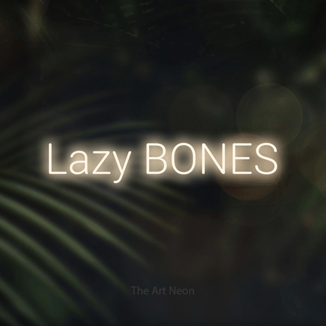 Warm white LED neon sign reading "Lazy Bones" against dark blurred palm leaves for bedroom décor