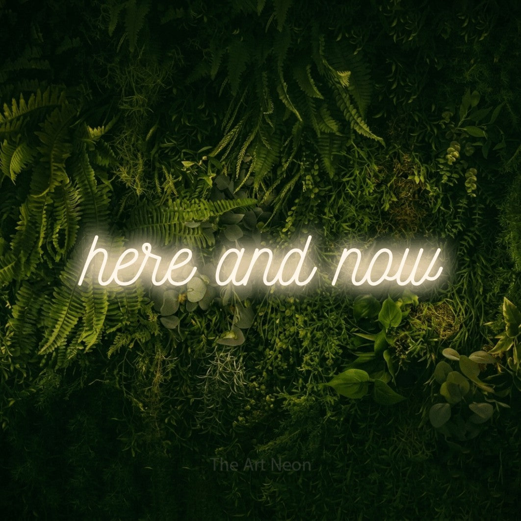Warm white script-style LED neon sign reading "Here and Now" against a moss-covered green wall for living room décor