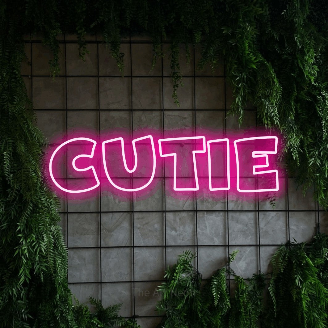 Enseigne néon à DEL à lumière rose vif, de style contour, affichant « Cutie » sur un mur quadrillé vert avec du feuillage, pour décorer une chambre de bébé