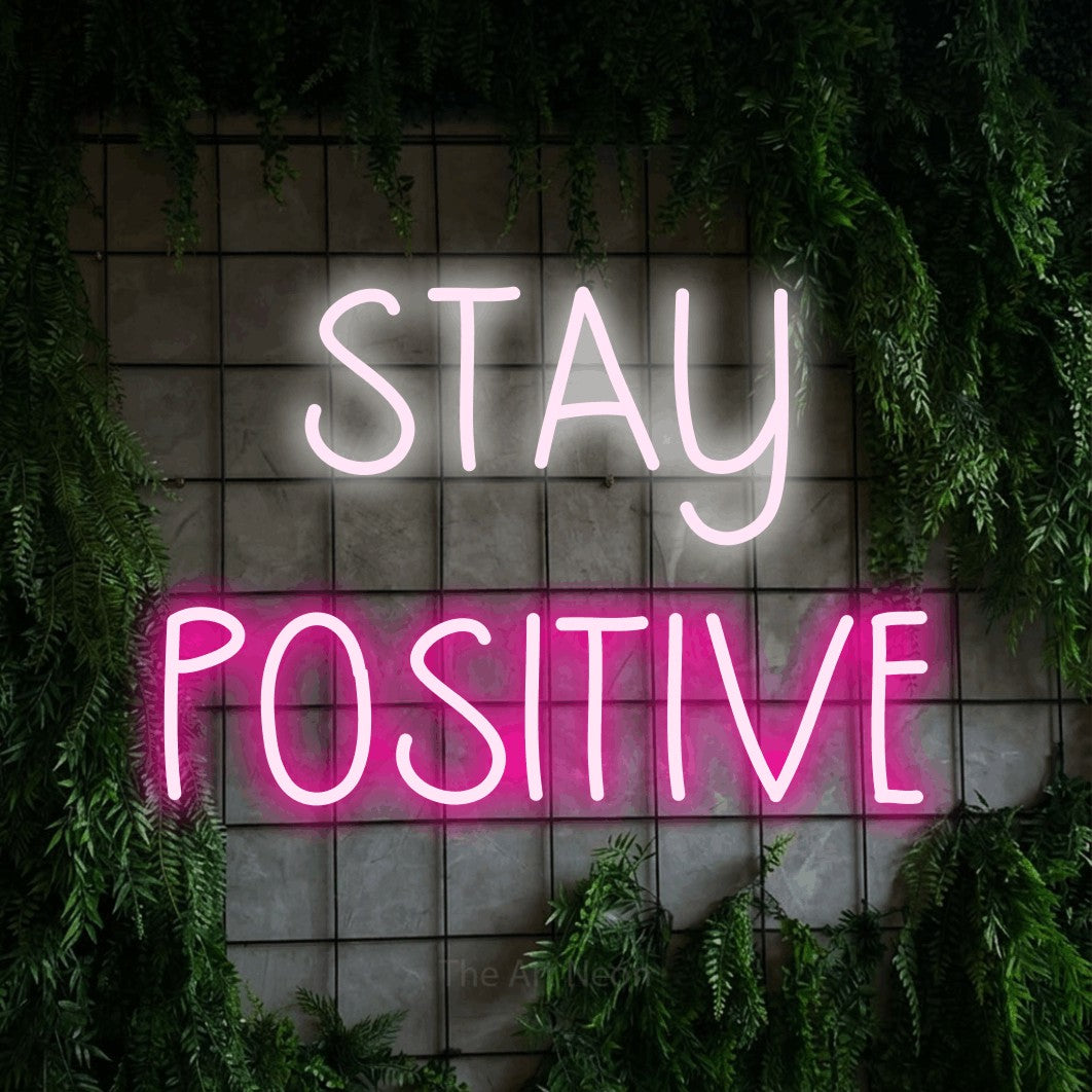 Hot pink and cool white LED neon sign reading "Stay Positive" against a green grid wall with foliage