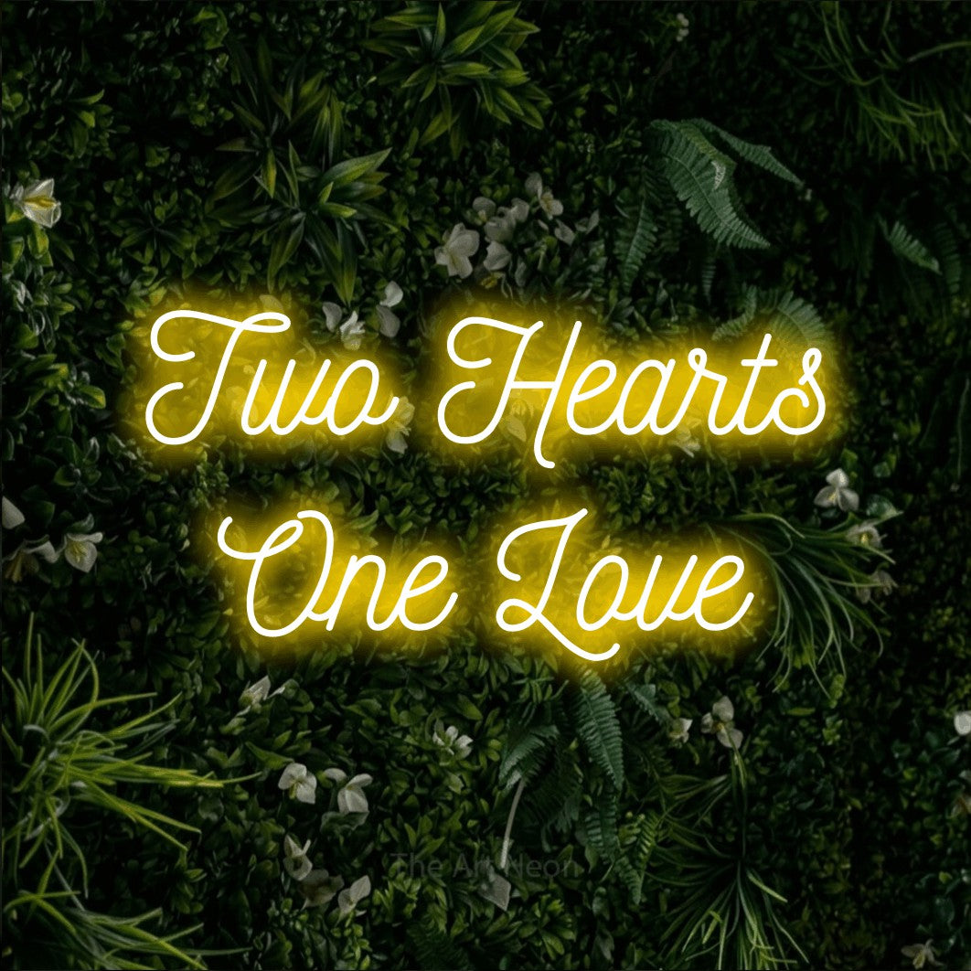 Yellow script-style LED neon sign reading "Two Hearts One Love" against a leafy green wall