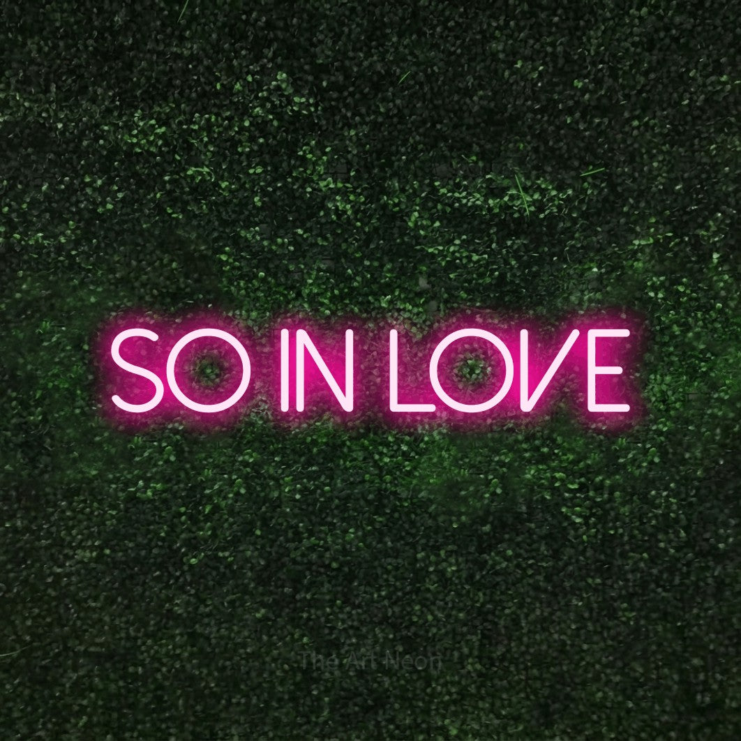 Hot pink LED neon sign reading "So in Love" against a textured green moss wall