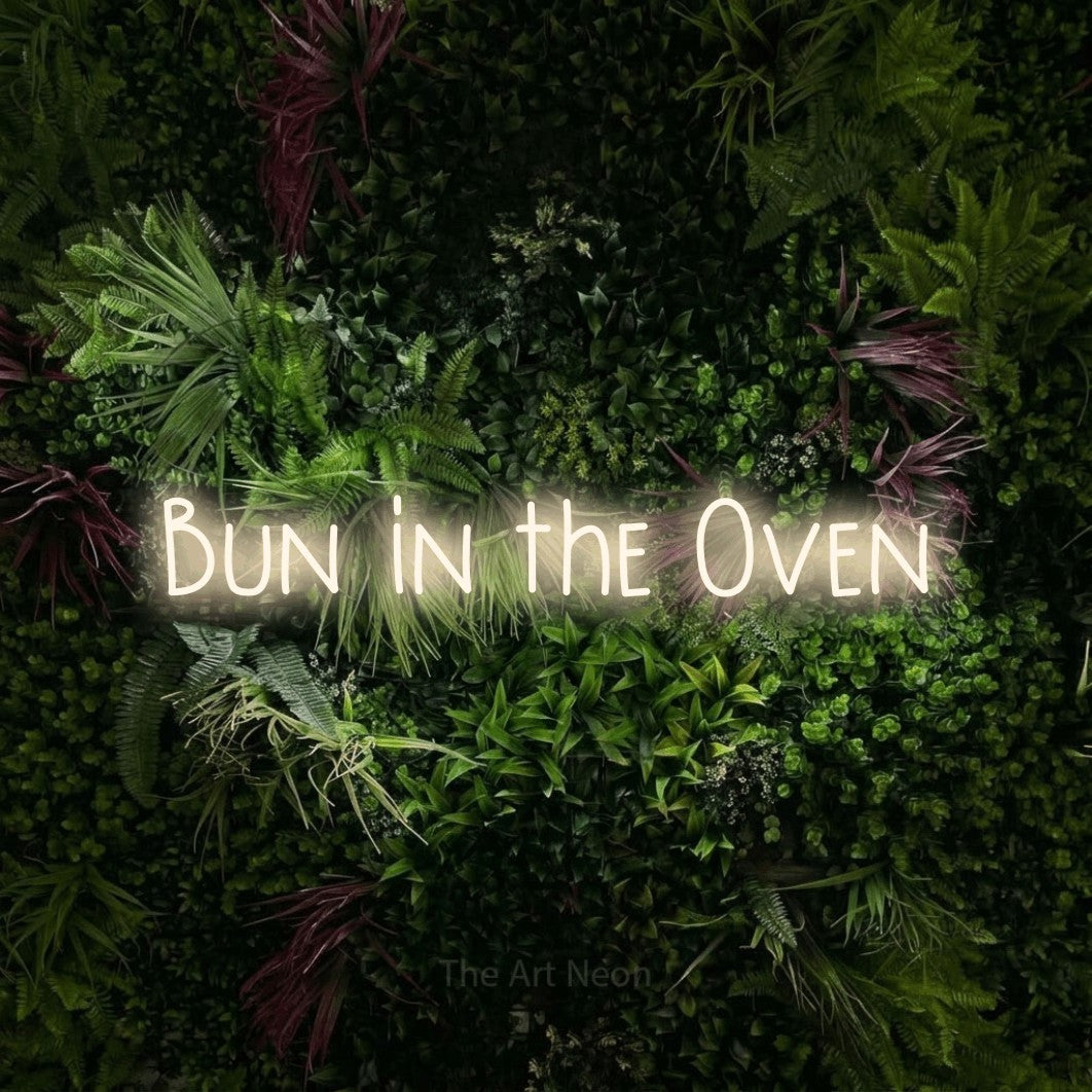 Warm white LED neon sign reading "Bun in the Oven" against a lush garden wall with pink flowers