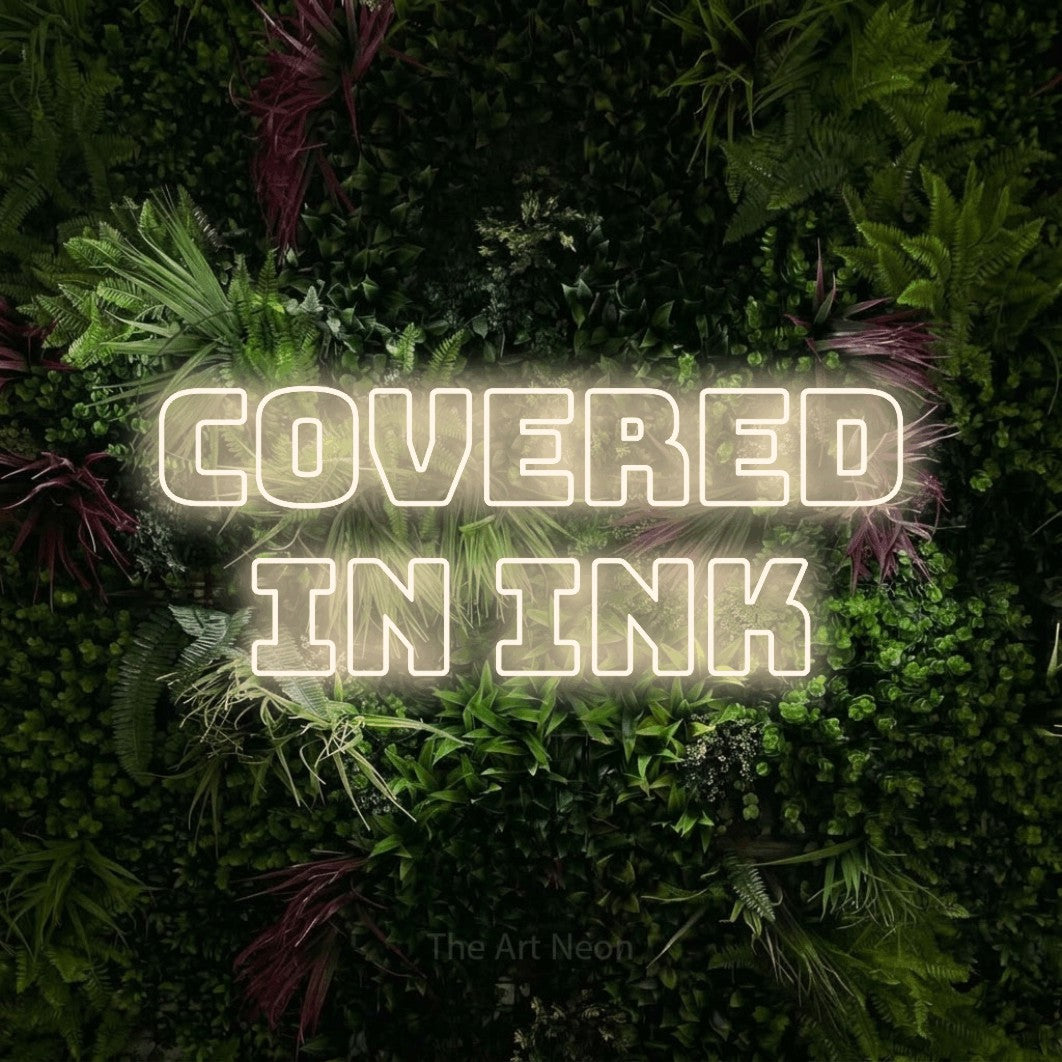 Warm white outline-style LED neon sign reading "Covered in Ink" against a lush garden wall with pink flowers