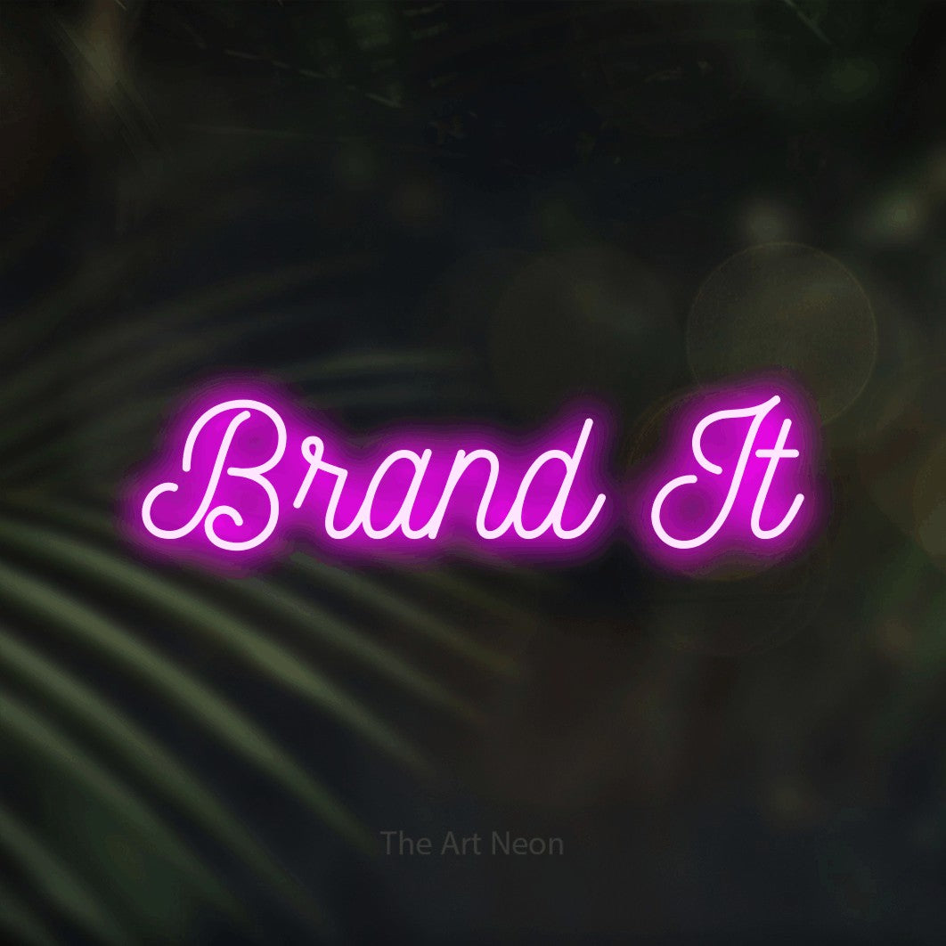 Pink script-style LED neon sign reading "Brand It" against dark blurred palm leaves for office décor