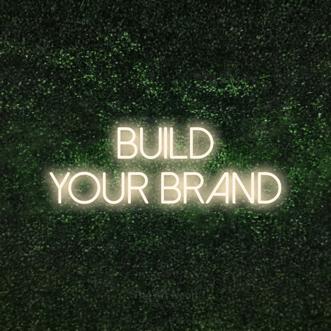 Warm white LED neon sign reading "Build Your Brand" against a textured green moss wall for office décor