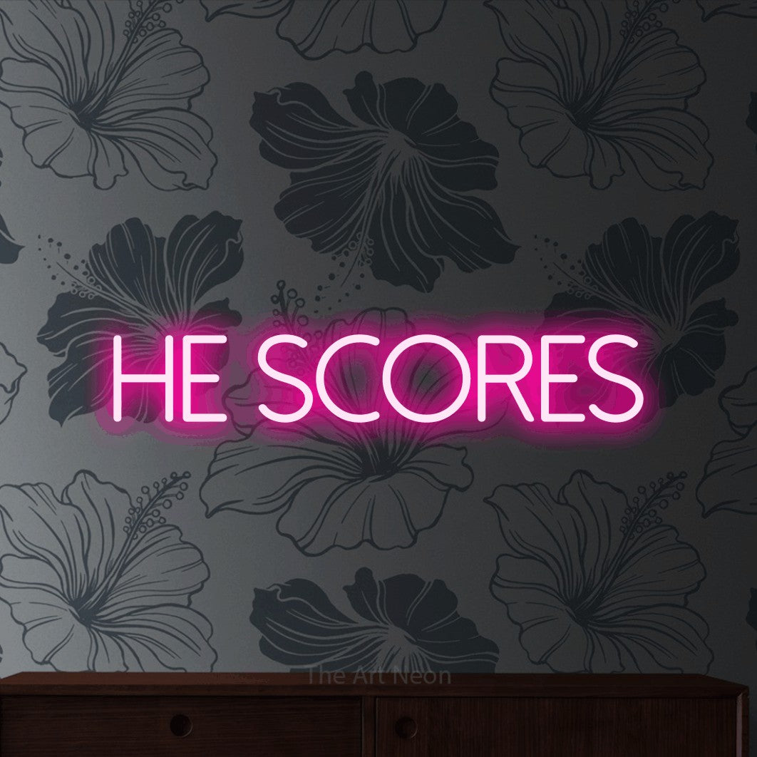 Hot pink LED neon sign reading "He Scores" against grey floral wallpaper