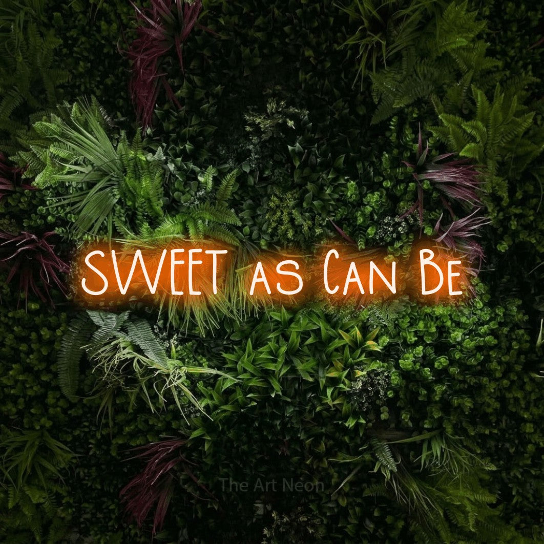 Orange LED neon sign reading "Sweet as Can Be" against a lush garden wall with pink flowers