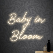 Warm white script-style LED neon sign reading "Baby in Bloom" against light grey floral wallpaper