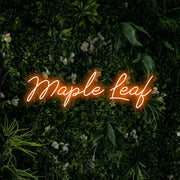 Orange script-style LED neon sign reading "Maple Leaf" against a leafy green wall