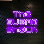 Purple outline-style LED neon sign reading "The Sugar Shack" against a dark geometric wood wall