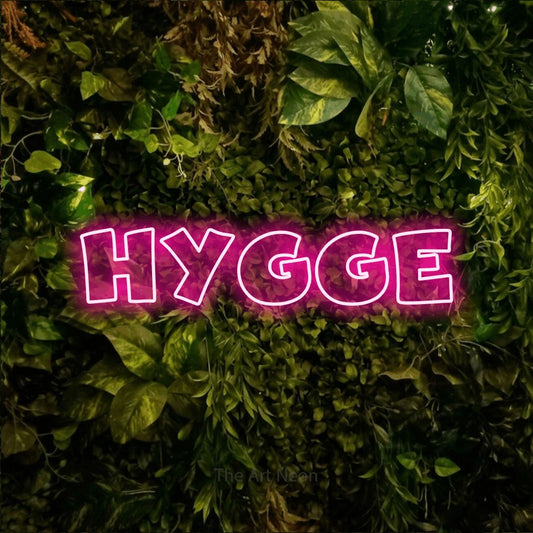 Hot pink outline-style LED neon sign reading "Hygge" against a textured moss and foliage wall for bedroom décor