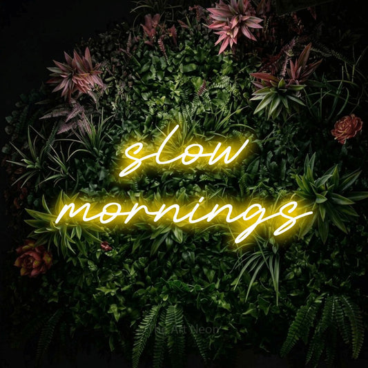 Yellow LED neon sign reading "Slow Mornings" against a flowering garden wall for bedroom décor
