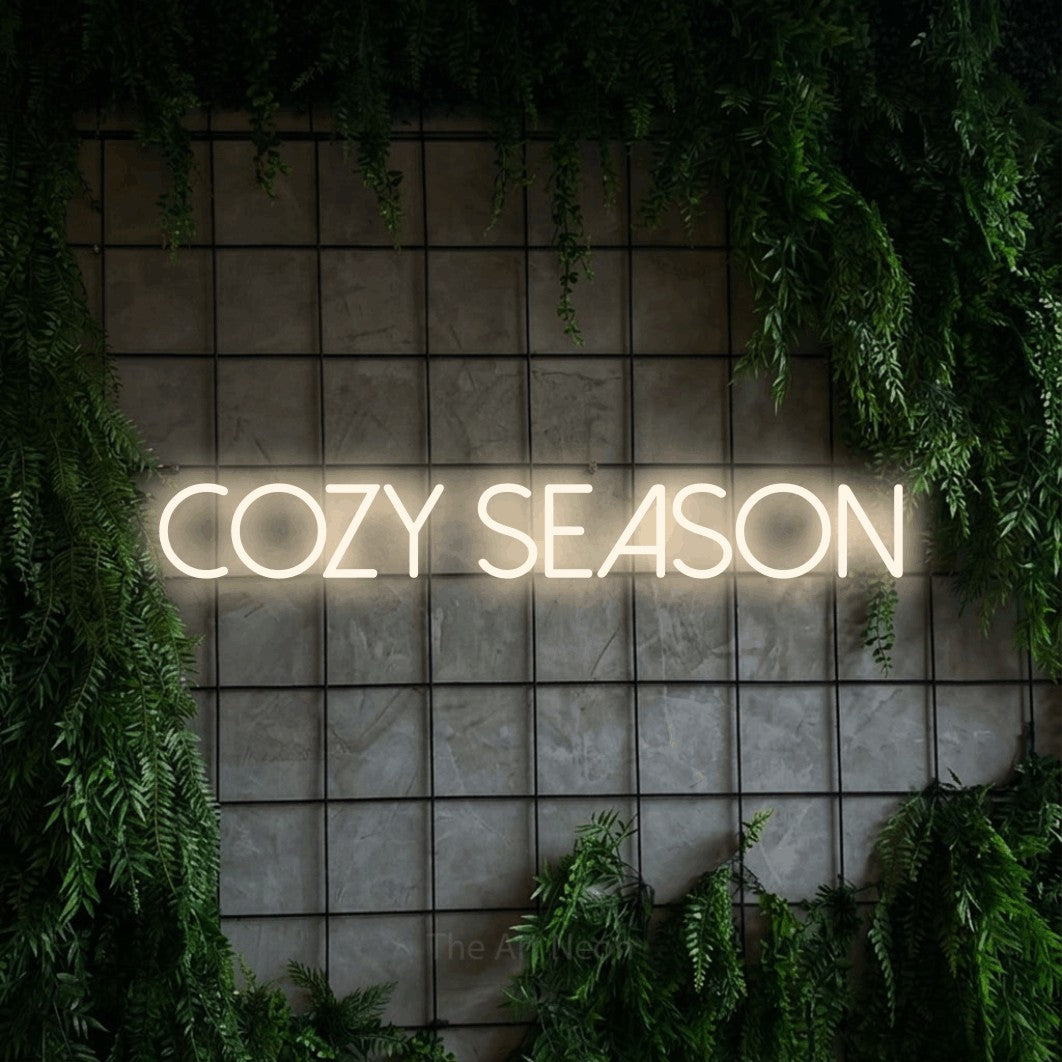 Warm white LED neon sign reading "Cozy Season" against a green grid wall with foliage for bedroom décor