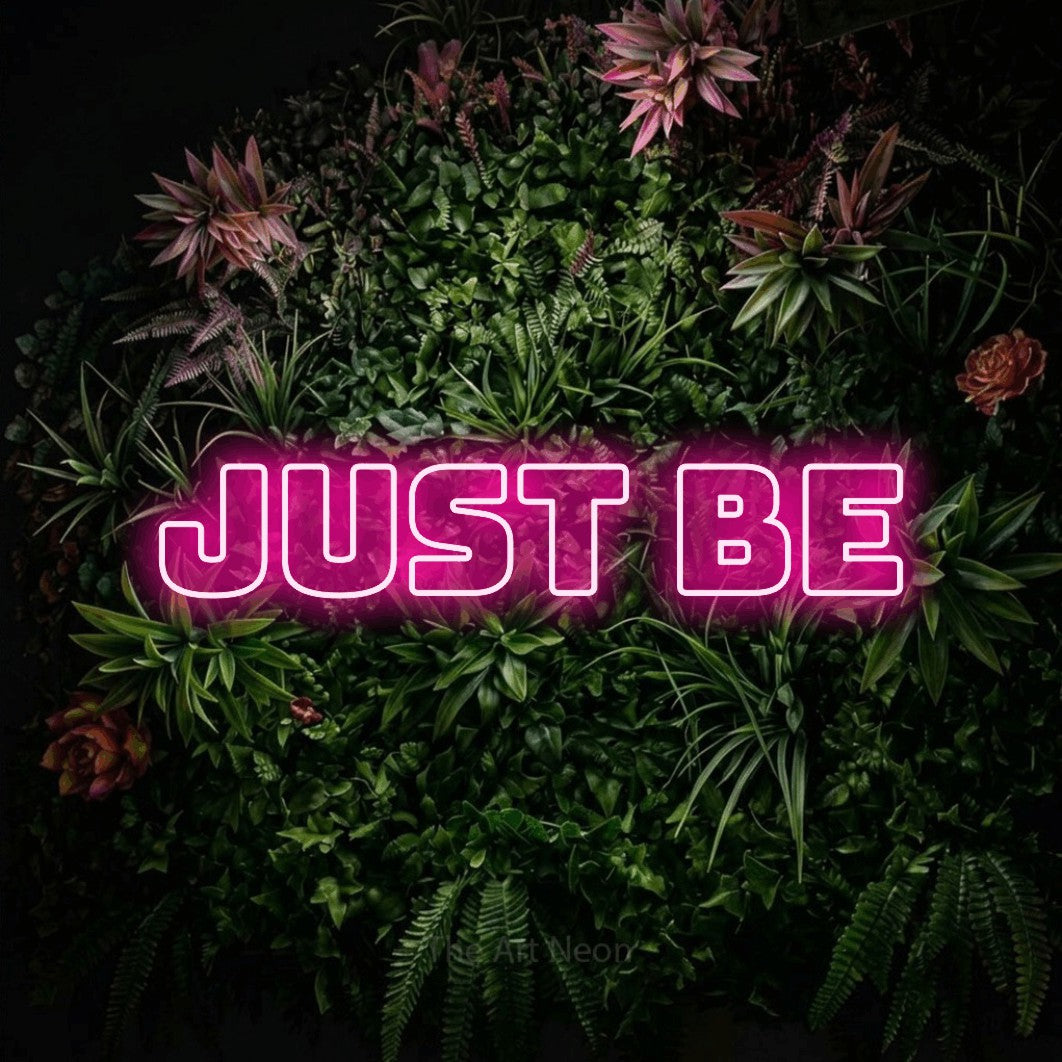 Hot pink outline-style LED neon sign reading "Just Be" against a flowering garden wall for bedroom décor