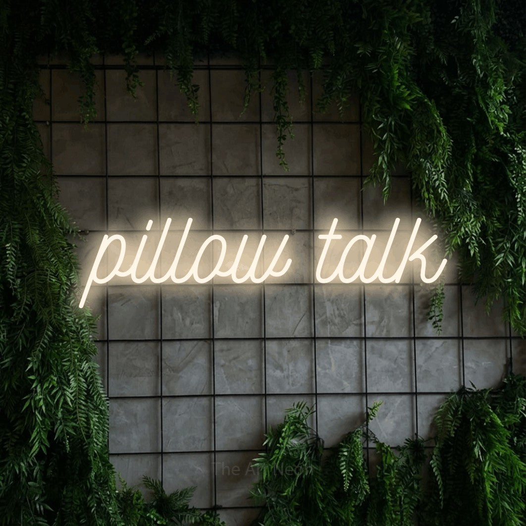 Warm white script-style LED neon sign reading "Pillow Talk" against a green grid wall with foliage for bedroom décor
