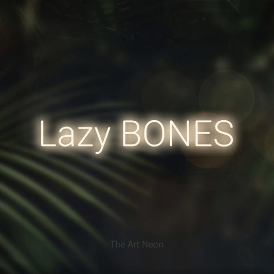 Warm white LED neon sign reading "Lazy Bones" against dark blurred palm leaves for bedroom décor