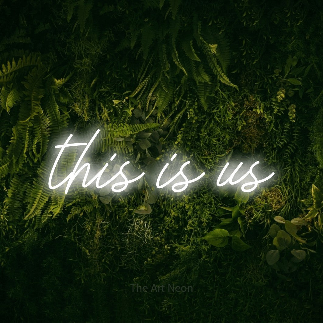 Cool white script-style LED neon sign reading "This is Us" against a moss-covered green wall for living room décor