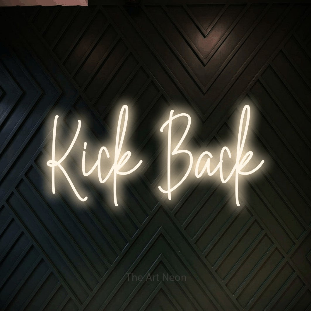 Warm white script-style LED neon sign reading "Kick Back" against a dark geometric wood wall for living room décor