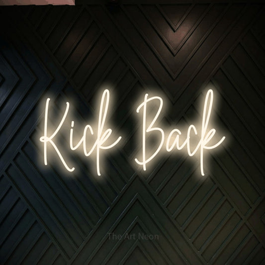 Warm white script-style LED neon sign reading "Kick Back" against a dark geometric wood wall for living room décor