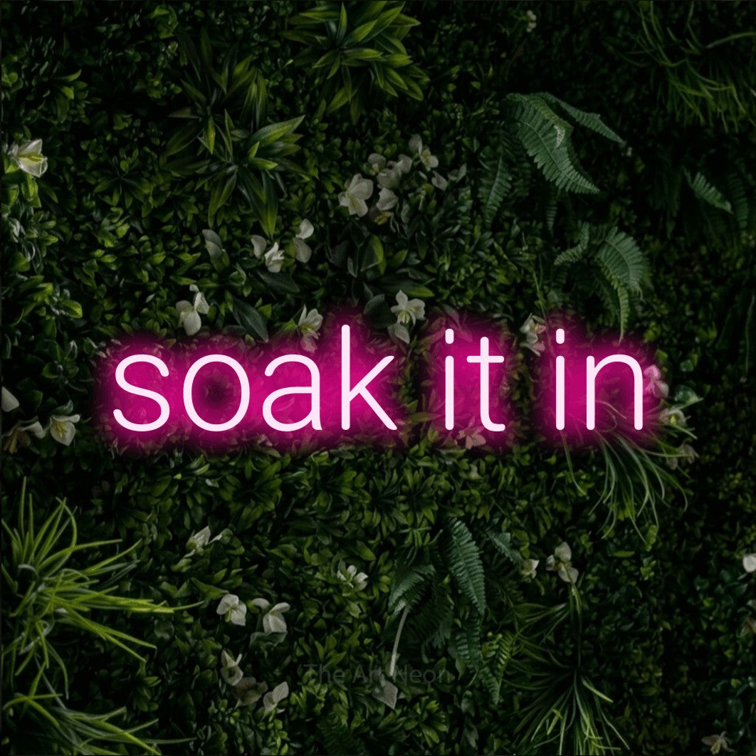 Hot pink LED neon sign reading "Soak It In" against a leafy green wall for living room décor