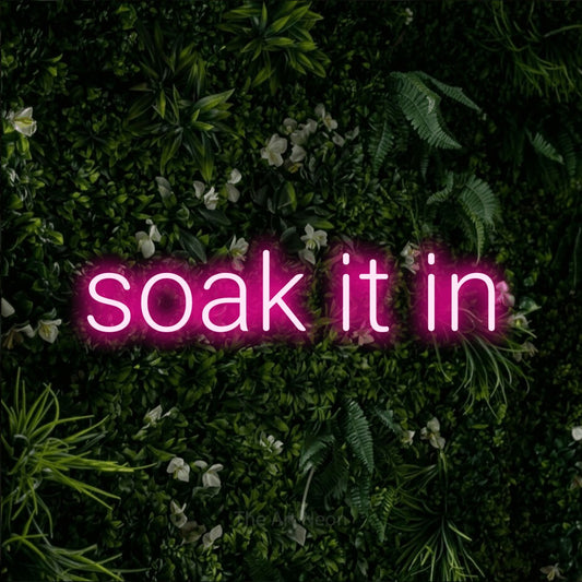 Hot pink LED neon sign reading "Soak It In" against a leafy green wall for living room décor