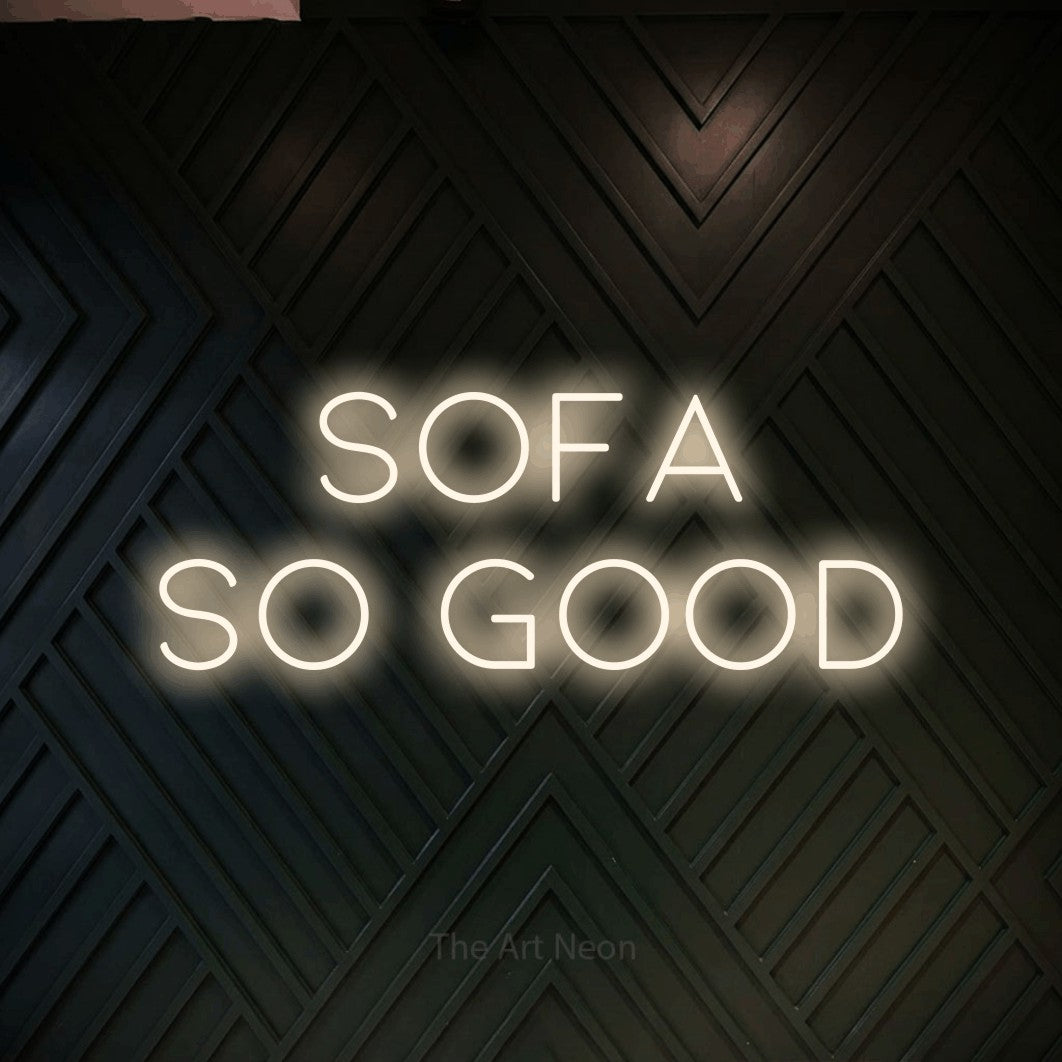 Warm white LED neon sign reading "Sofa So Good" against a dark geometric wood wall for living room décor