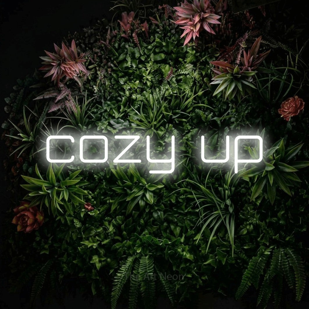 Cool white outline-style LED neon sign reading "Cozy Up" against a flowering garden wall for living room décor