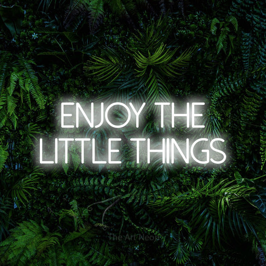 Cool white LED neon sign reading "Enjoy the Little Things" against a dense tropical foliage wall for living room décor