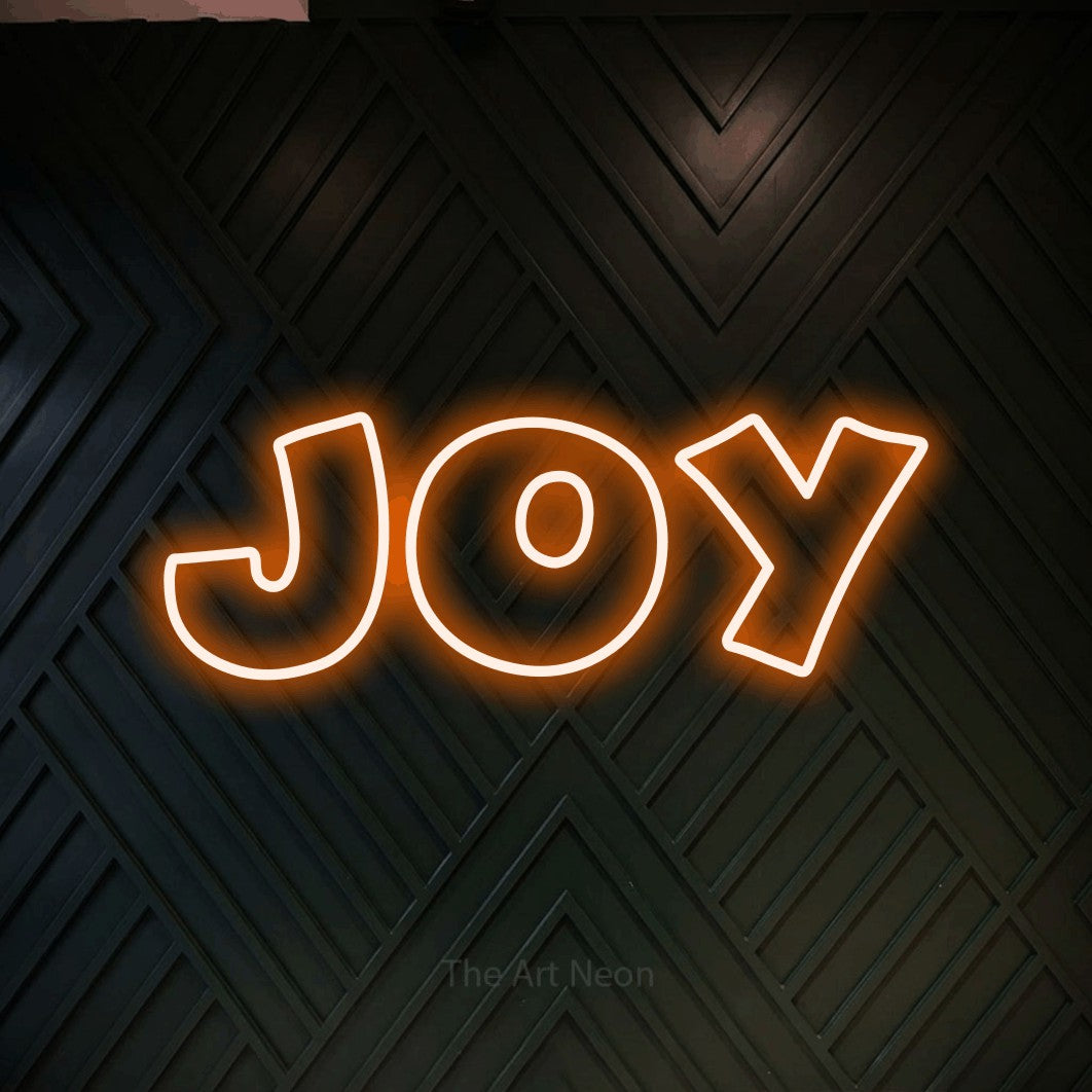 Orange outline-style LED neon sign reading "Joy" against a dark geometric wood wall for living room décor