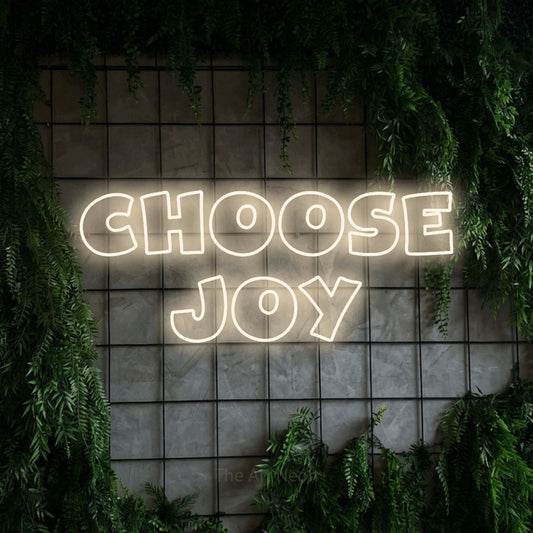 Warm white outline-style LED neon sign reading "Choose Joy" against a green grid wall with foliage for living room décor