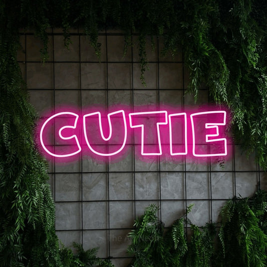 Hot pink outline-style LED neon sign reading "Cutie" against a green grid wall with foliage for nursery décor