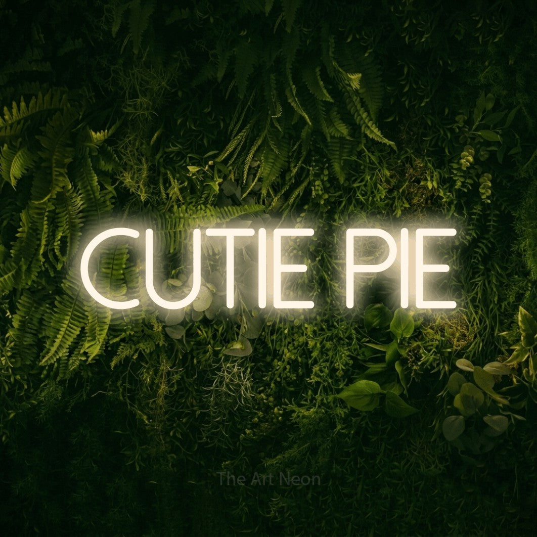 Warm white LED neon sign reading "Cutie Pie" against a moss-covered green wall for nursery décor