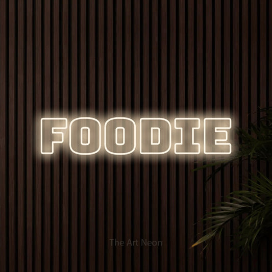 Warm white outline-style LED neon sign reading "Foodie" against dark wooden slats for kitchen décor