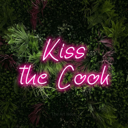 Hot pink LED neon sign reading "Kiss the Cook" against a lush garden wall with pink flowers for kitchen décor