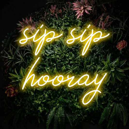 Yellow script-style LED neon sign reading "Sip Sip Hooray" against a flowering garden wall for kitchen décor