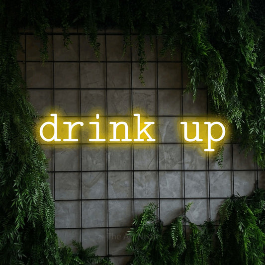 Yellow LED neon sign reading "Drink Up" against a green grid wall with foliage for kitchen décor