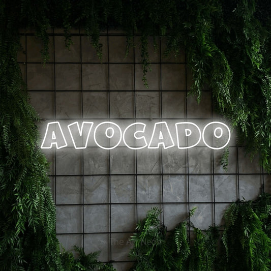Cool white outline-style LED neon sign reading "Avocado" against a green grid wall with foliage for kitchen décor