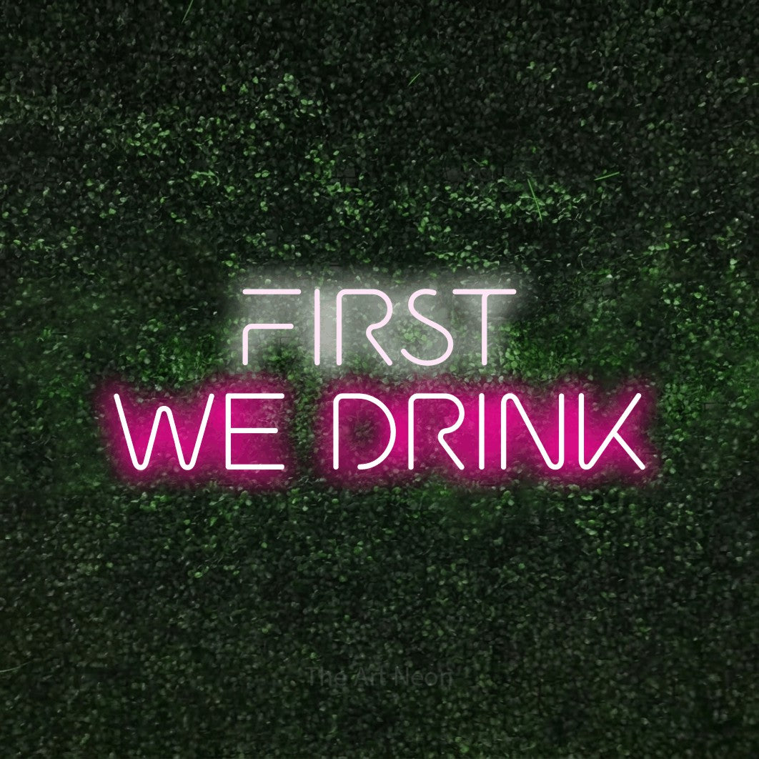 Hot pink and cool white LED neon sign reading "First We Drink" against a textured green moss wall for bar décor