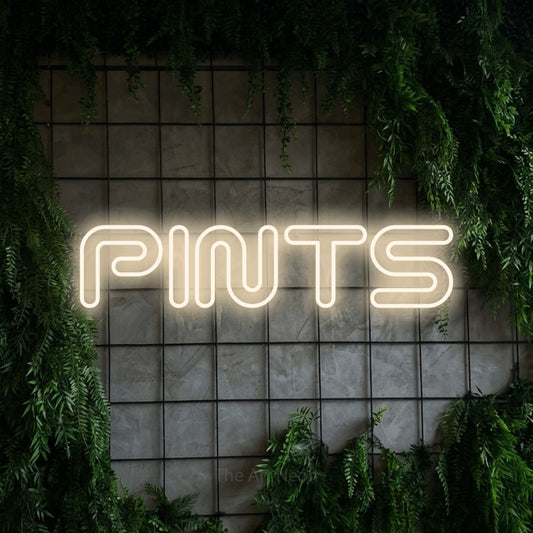 Warm white outline-style LED neon sign reading "Pints" against a green grid wall with foliage for bar décor