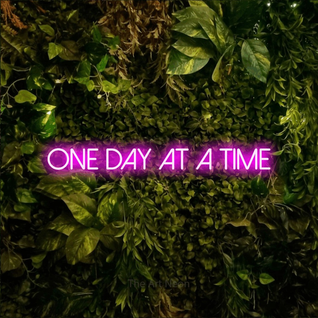 Pink LED neon sign reading "One Day at a Time" against a textured moss and foliage wall