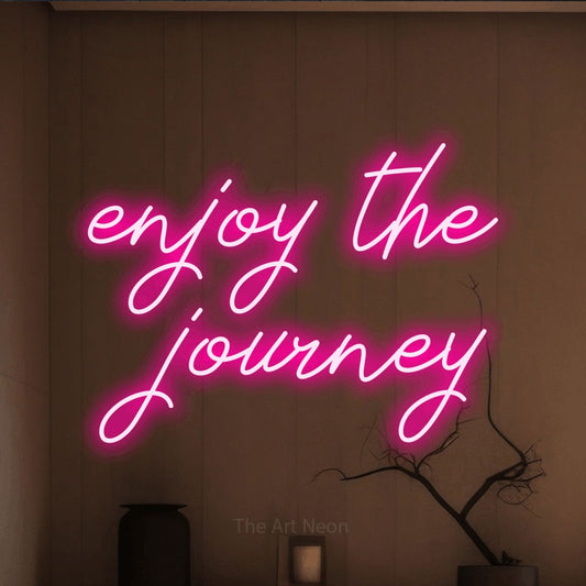 Hot pink script-style LED neon sign reading "Enjoy the Journey" against a warm brown wall