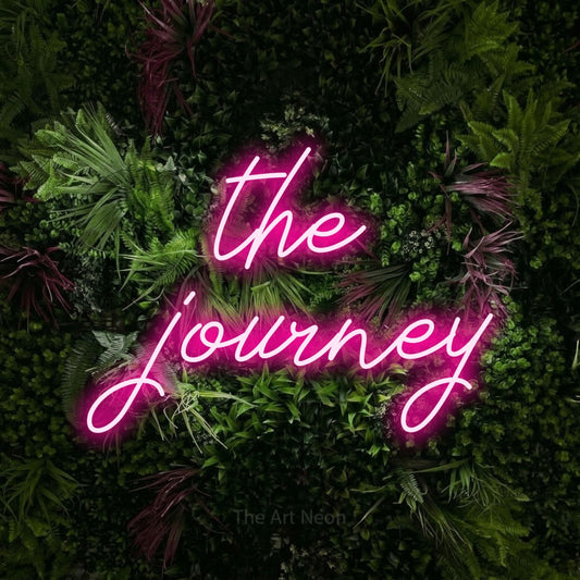 Hot pink script-style LED neon sign reading "The Journey" against a lush garden wall with pink flowers