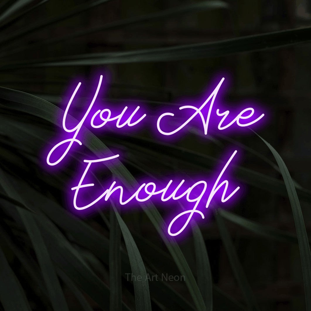 Purple script-style LED neon sign reading "You Are Enough" against dark tropical leaves