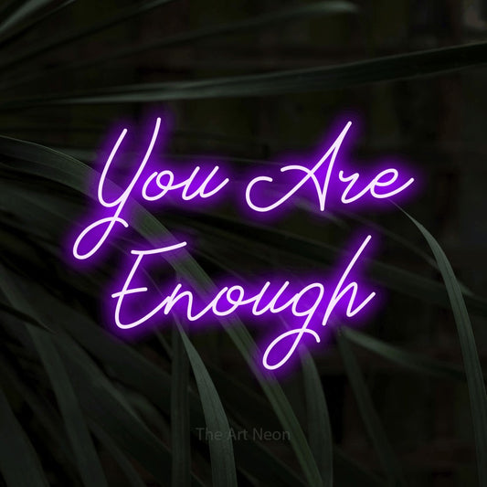 Purple script-style LED neon sign reading "You Are Enough" against dark tropical leaves