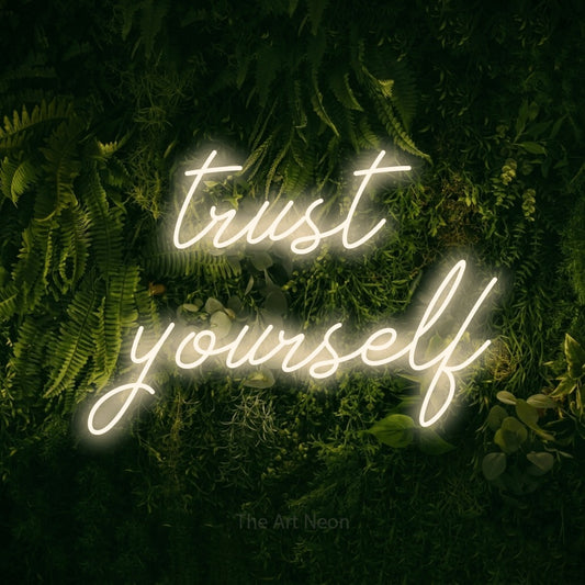 Warm white script-style LED neon sign reading "Trust Yourself" against a moss-covered green wall