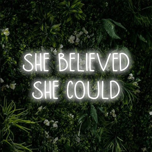 Cool white LED neon sign reading "She Believed She Could" against a leafy green wall