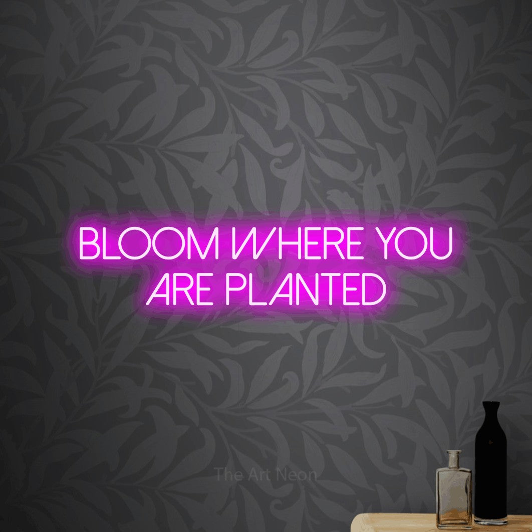 Pink LED neon sign reading "Bloom Where You Are Planted" against light grey floral wallpaper