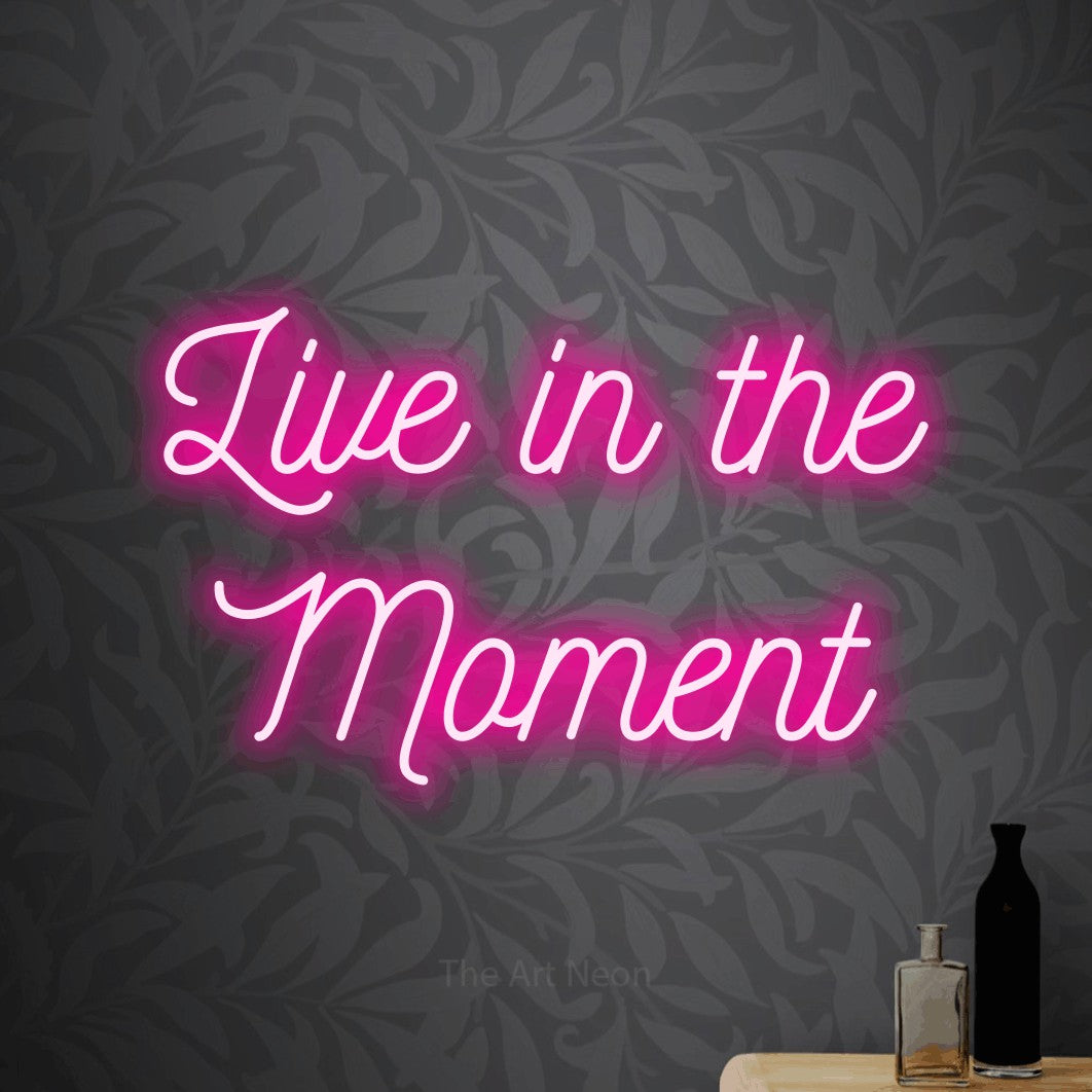 Hot pink script-style LED neon sign reading "Live in the Moment" against light grey floral wallpaper