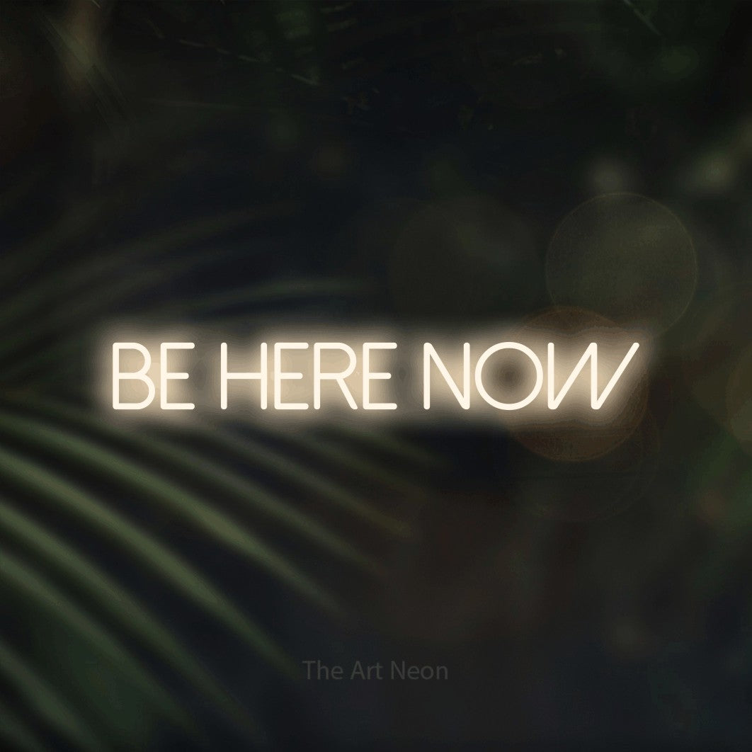 Warm white LED neon sign reading "Be Here Now" against dark blurred palm leaves